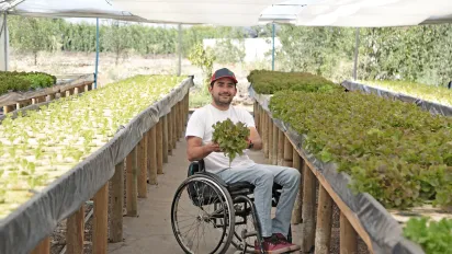Man in a wheelchair inside a greenhouse. He holds a lettuce in his hands and smiles at the camera.