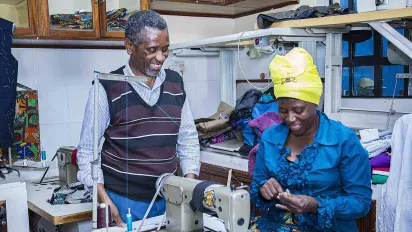 A portrait of Celestin Mbaruku. He stands beside a trainee in a sewing workshop. The trainee and teacher both smile broadly. (Nairobi, Kenya 2024)