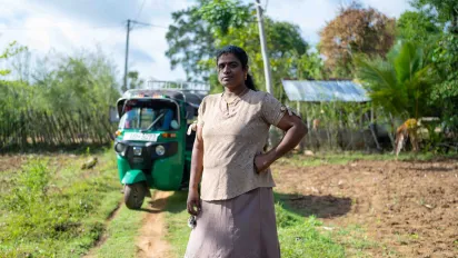 A portrait of Leena Penildas Thatkuras. She stands on a path between two fields, with her hand on her hip. Her three-wheeler is parked in the background. (2024)