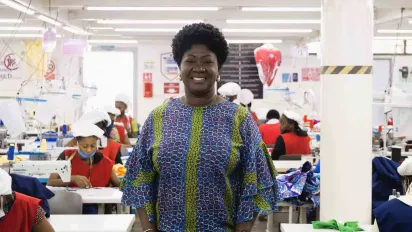 A portrait of Linda Ampah. She stands on the factory floor, between rows of garment workers at KAD Manufacturing Ltd in Accra, Ghana. (2024)