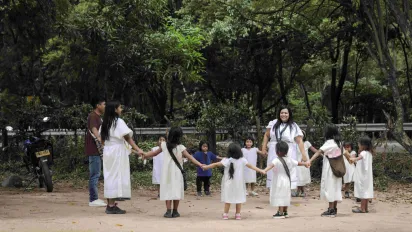 Indigenous woman Yoraima Cristina Navarro stands in a circle of children and adults holding hands. They are outside and there are leafy green trees in the background. (2025)