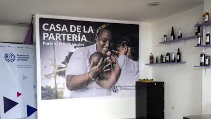 Interior de la nueva Casa de la Partería en Quibdó. En la pared se ve un gran afiche con la imagen de una partera afrodescendiente sonriendo mientras carga a un bebé, acompañado del texto “Casa de la Partería – Parter@s tradicionales” y el logo de ASOREDIPAR. A la izquierda hay un pendón de la OIT y a la derecha estantes con frascos de preparados medicinales.