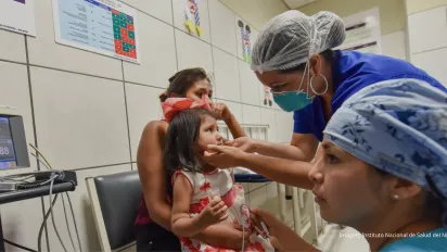Niña pequeña en vestido rojo sentada en el regazo de su madre mientras una trabajadora de salud le revisa la boca en un consultorio pediátrico.