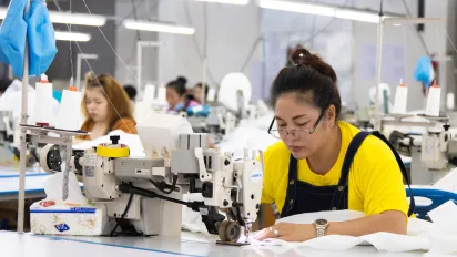 A woman sewing, with sewing machines and other seamstresses working in the background.