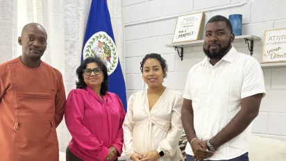 A group portrait of four people standing indoors in front of a Belize flag, smiling at the camera