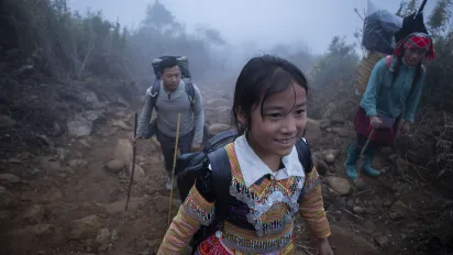 A young ethnic minority girl walks uphill on a rocky, fog-covered mountain trail, carrying a backpack, with adult trekkers following behind her.