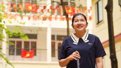 A young Vietnamese woman with a visual impairment stands smiling in a school courtyard, holding a white cane.