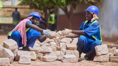 Smiling young workers pass blocks hand in hand as part of labour‑intensive road rehabilitation works.