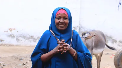 Portrait of Hawa Ali Mohamed. She stands with her hands clasped and wears a blue hijab and abaya. A cow stands behind her in the background.
