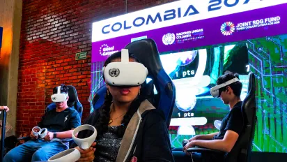 Tres personas participan en una experiencia de realidad virtual sentadas en sillas tipo simulador. En primer plano, una joven sostiene los controles de un visor VR blanco con el emblema de las Naciones Unidas. Al fondo, una gran pantalla iluminada muestra el título “COLOMBIA 2077” junto con logotipos de Naciones Unidas y del Fondo Conjunto para los ODS. El espacio tiene paredes de ladrillo visto y una ambientación tecnológica con luces de colores.