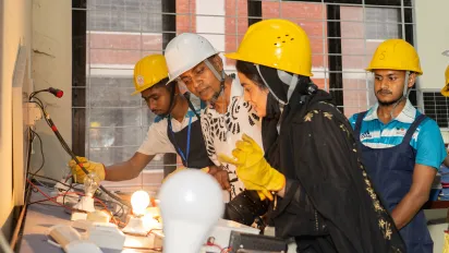 Vocational training institute principal Md. Ziaur Rahman and four trainees, including a female trainee in the foreground, wear hard hats and safety gloves and look at an electrical installation with two lit lightbulbs.