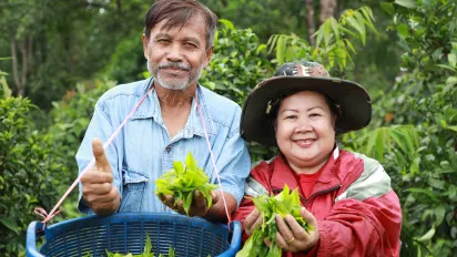 Two farmers stand in a tea plantation, smiling at the camera while holding freshly picked tea leaves.