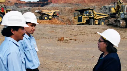Three people wearing white hard hats stand in conversation at an open-pit mining site.