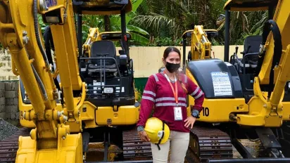 A woman wearing a high-visibility work shirt, face mask, and identification badge stands between two mini excavators while holding a yellow safety helmet.
