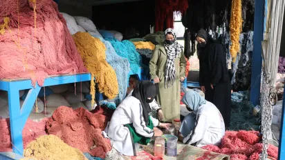 Women work among large bundles of colourful yarn while two other women observe their work.