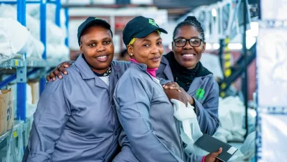 Three women factory workers in grey uniforms smile together during an ILO project field visit in South Africa in 2023.