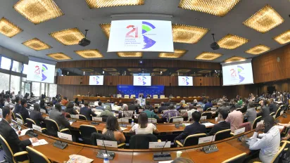 Wide-angle view of delegates seated around curved conference tables, facing a panel of speakers, with screens displaying the conference logo