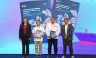 Four people standing on the stage and two of them holding boxes containing a publication
