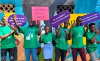 A group of young people in green T-shirts stand in front of a colourful mural, smiling and holding signs calling for innovations to end child labour in their communities.