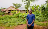 A young Nigerian man, David Chukwuemeka, stands smiling in front of his family’s rural home in Aponmu, Ondo State. He is wearing a blue shirt, surrounded by green plants and simple houses in the background.