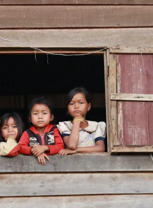 Three children are peeking out of a window in a wooden house.