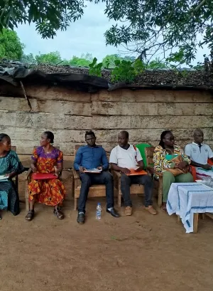 in a community, people seat in chairs under a tree with a table in the front, they seat in front of a house built with handmade bricks 