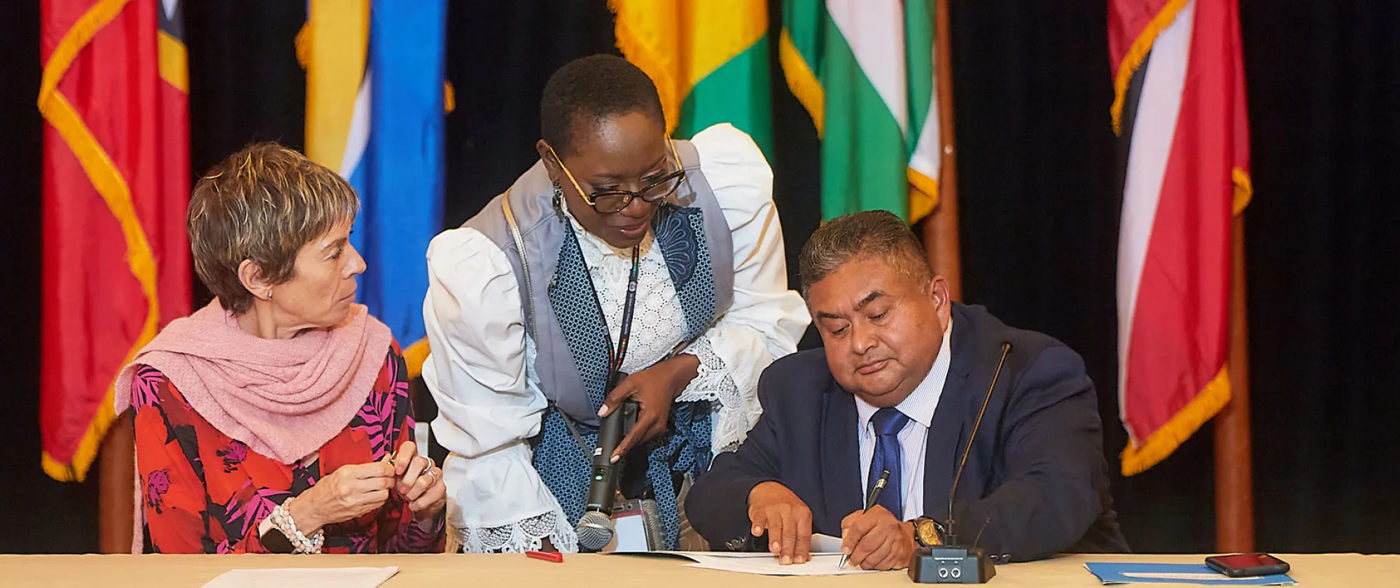 A man signing a document sitting next to a woman with a woman standing behind them