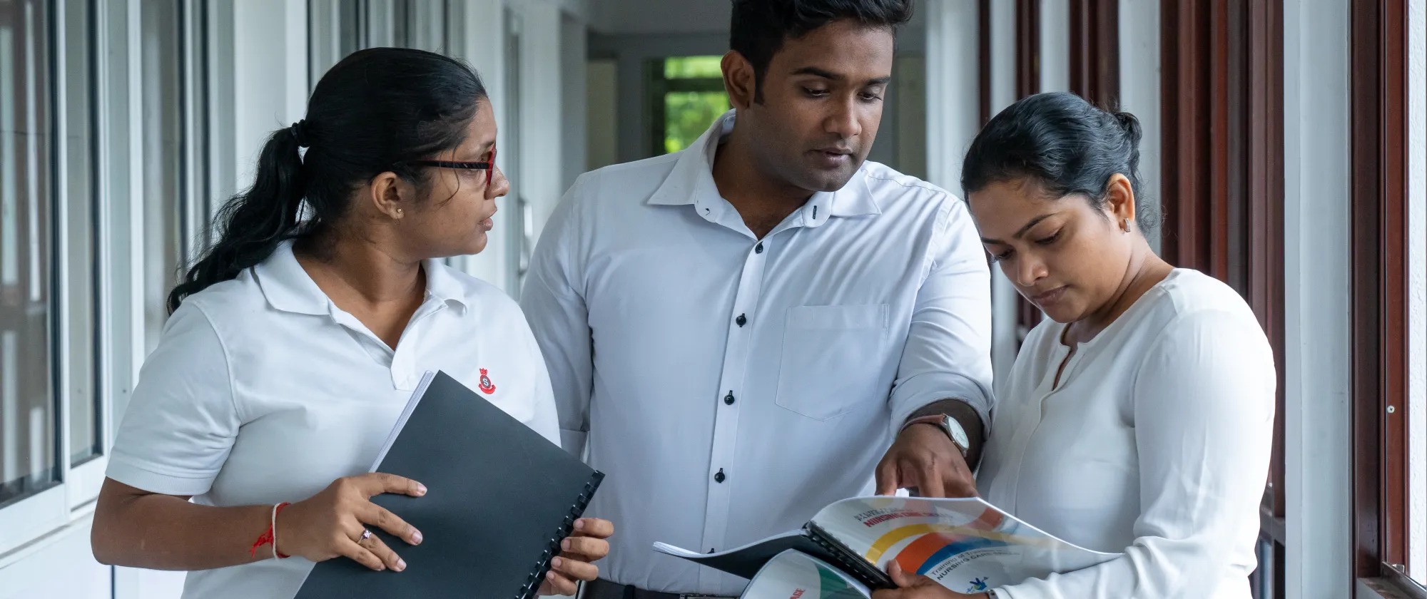 A man and 2 women walk along a corridor while examining a training manual book 
