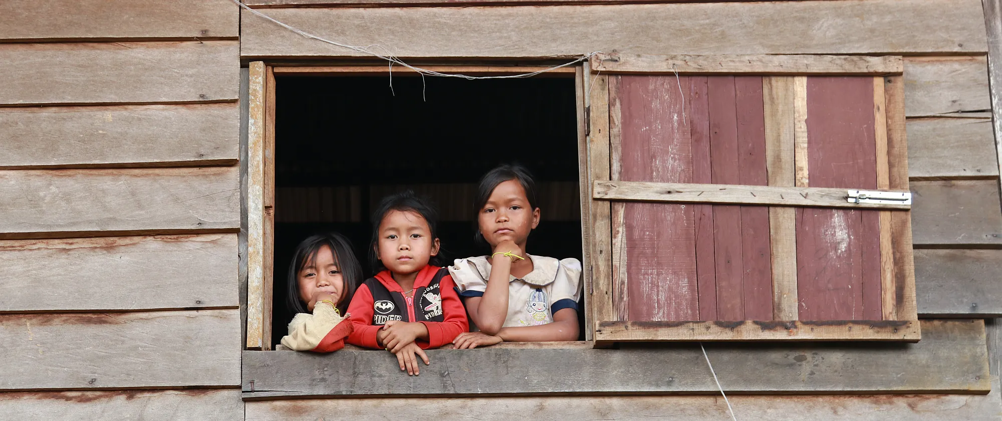 Three children are peeking out of a window in a wooden house.