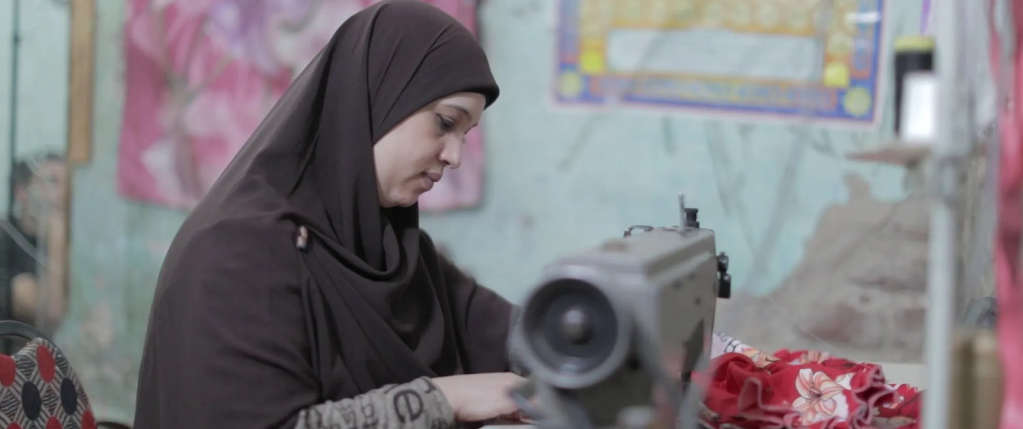 A woman wearing a headscarf sews fabric on a sewing machine in a room with colourful posters and fabric decorations on the walls.