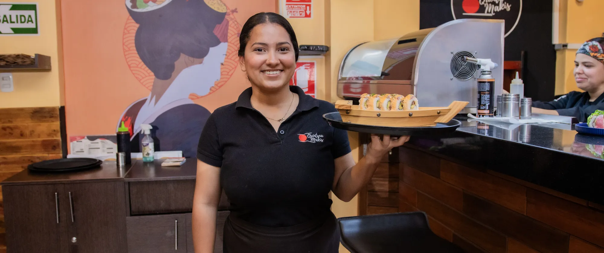 Mujer joven posa con bandeja en mano ante la barra de un restaurante
