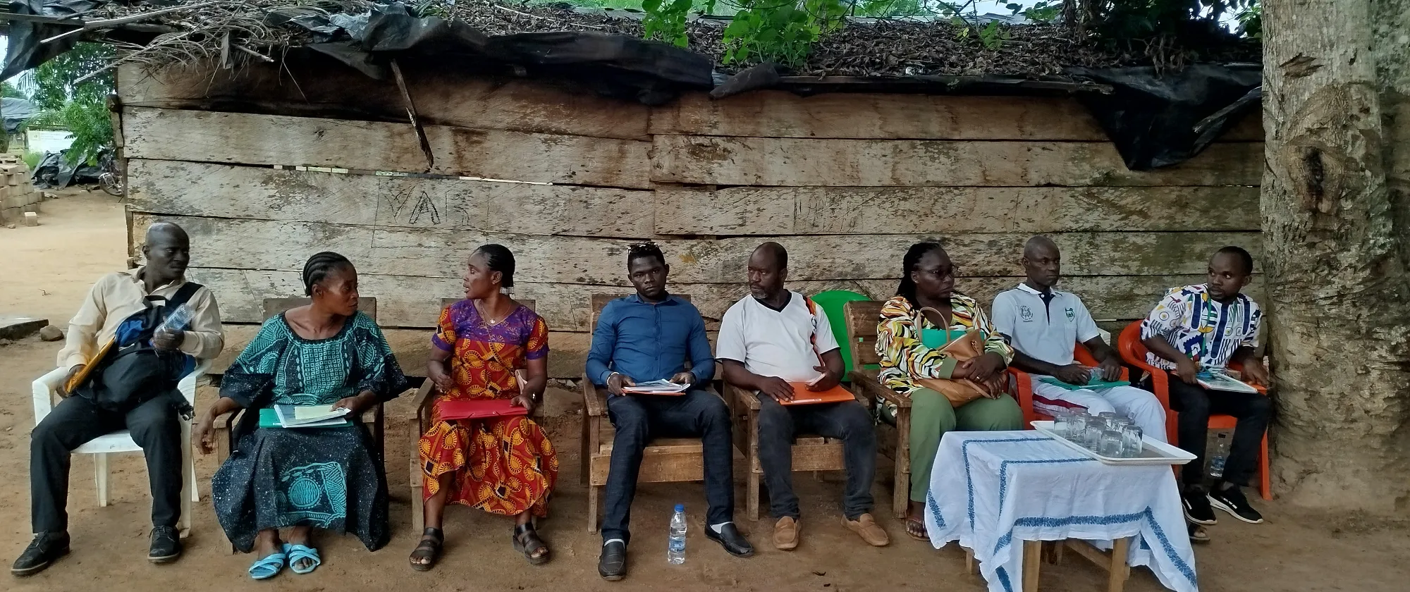 in a community, people seat in chairs under a tree with a table in the front, they seat in front of a house built with handmade bricks 
