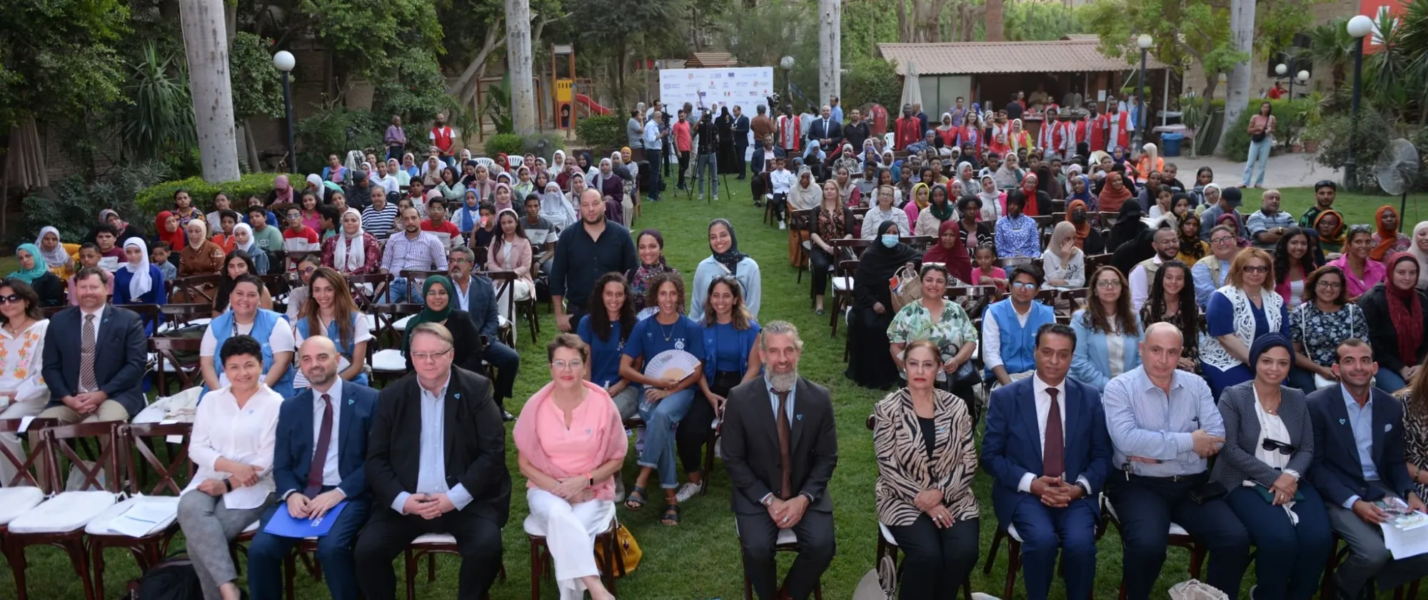 A diverse crowd of individuals seated on chairs in a grassy outdoor area.
