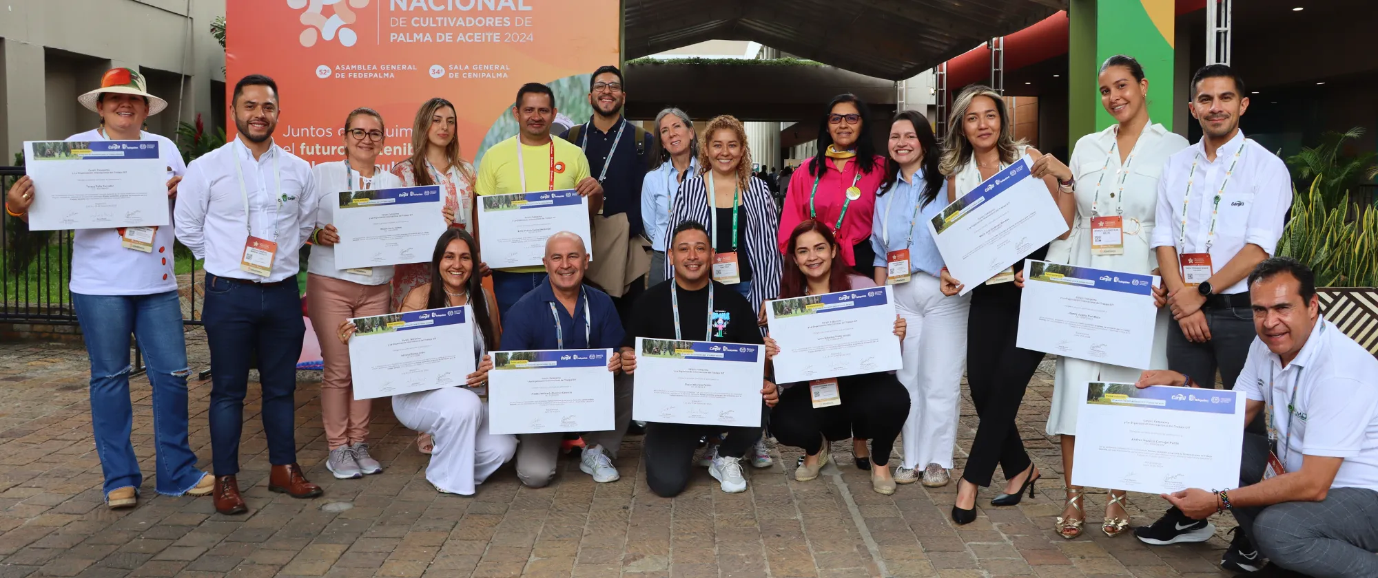 Foto grupal de los mejores estudiantes del diplomado Palma sostenible: Programa de formación para el trabajo decente en frente de un stand en el Congreso Palmero sosteniendo sus certificaciones