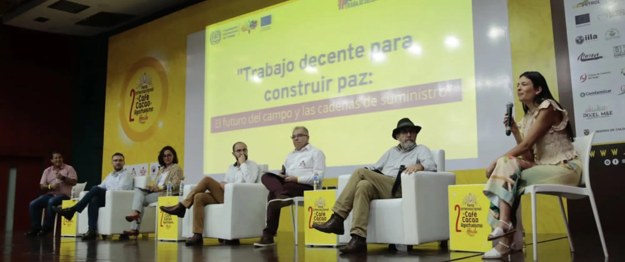De izquierda a derecha, están sentados los representantes de: la CUT, la OIT, FAO, Ministerio del Trabajo, la Federación Nacional de Cafeteros, Dignidad Agropecuaria y la moderadora del panel, Ángela Herrera. De fondo se muestra el nombre del espacio “Trabajo decente para construir paz: El futuro del campo y las cadenas de suministro”.