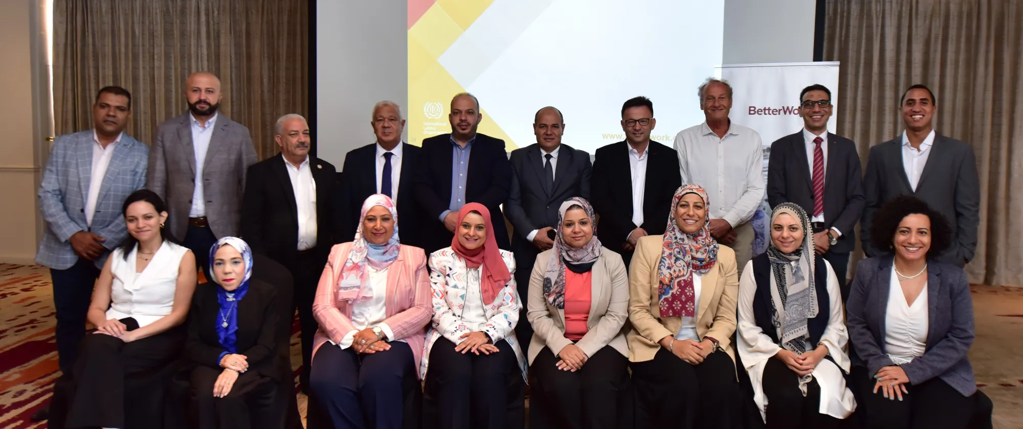 A group of people, dressed in business attire, pose for a photo in front of a screen displaying the words "Better Work".