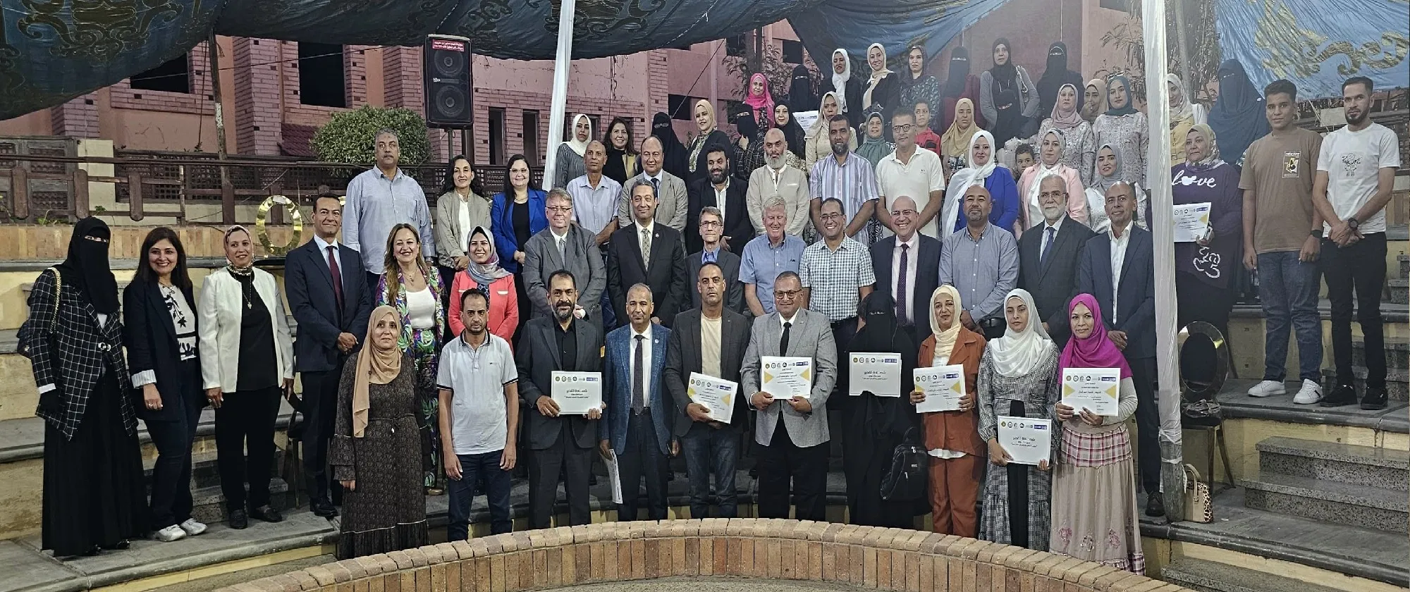 A large group of people, both men and women, pose for a photo outdoors. Some hold certificates. They stand on steps with a building and greenery in the background.
