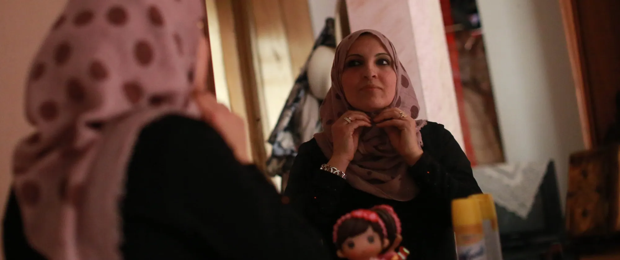 a veiled Palestinian woman fixing her veil while standing in front of a mirror as she gets ready to go to work.