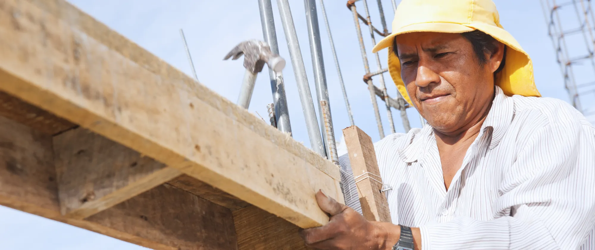 A photo of a man with a yellow building helmet is working on with a hammer on a wood structure