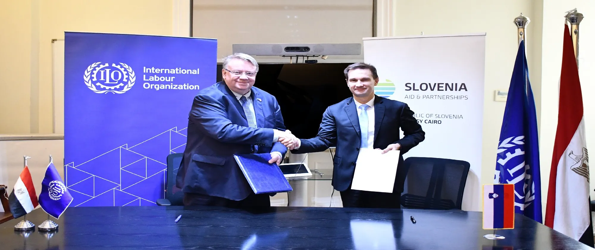 Two individuals shaking hands at an event with ILO and Slovenia partnership banners.