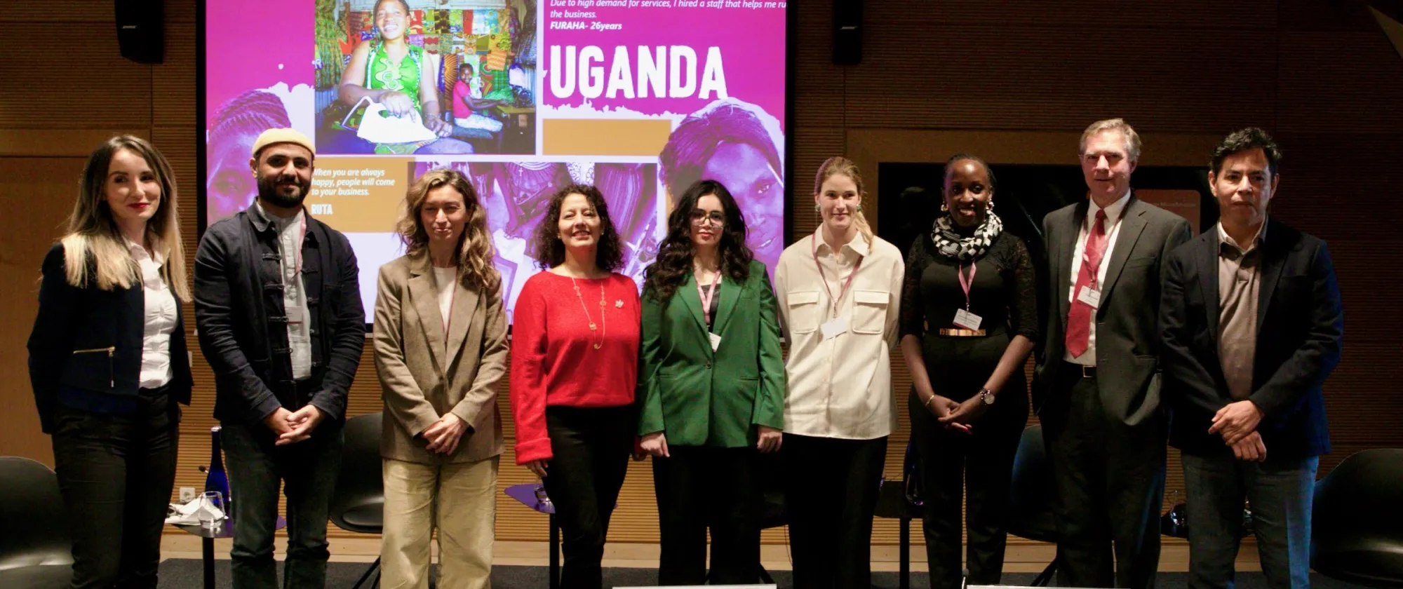 Nine individuals are side by side posing for a photo in front of a screen showing the name Uganda in a pink background
