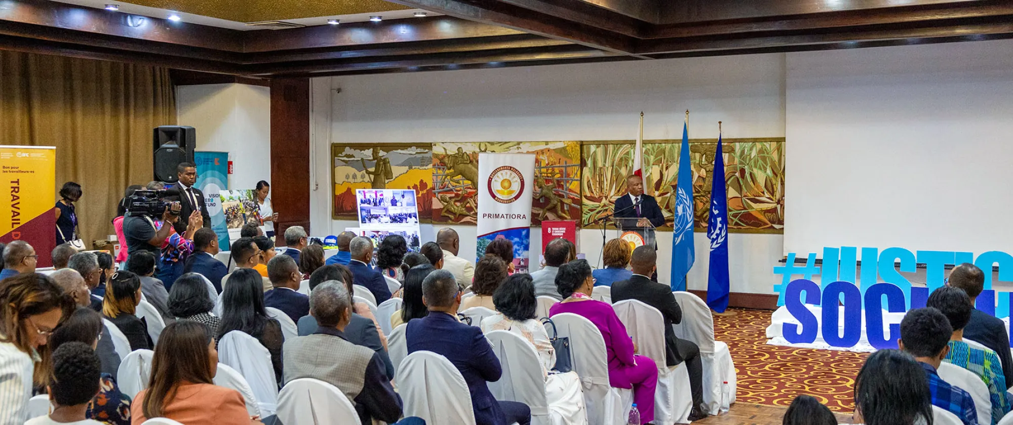 Une salle remplie de participants assis, écoutant un intervenant debout à un podium portant le logo de la Primature malgache. En arrière-plan, des drapeaux de l'ONU et de l'OIT, ainsi que des panneaux présentant des initiatives et projets liés au travail décent. À gauche, des bannières affichant des slogans sur le travail et la justice sociale.