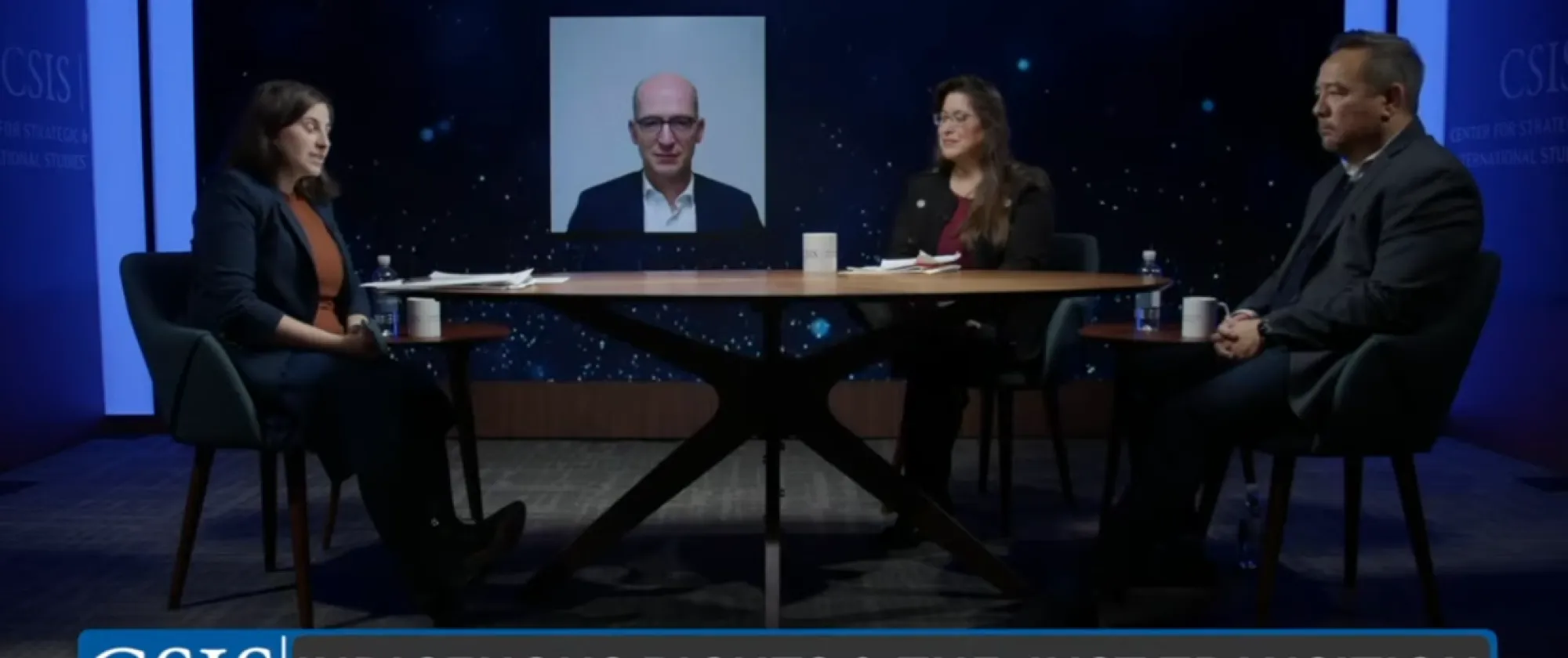 2 women and a man sitting at a table and one man on screen