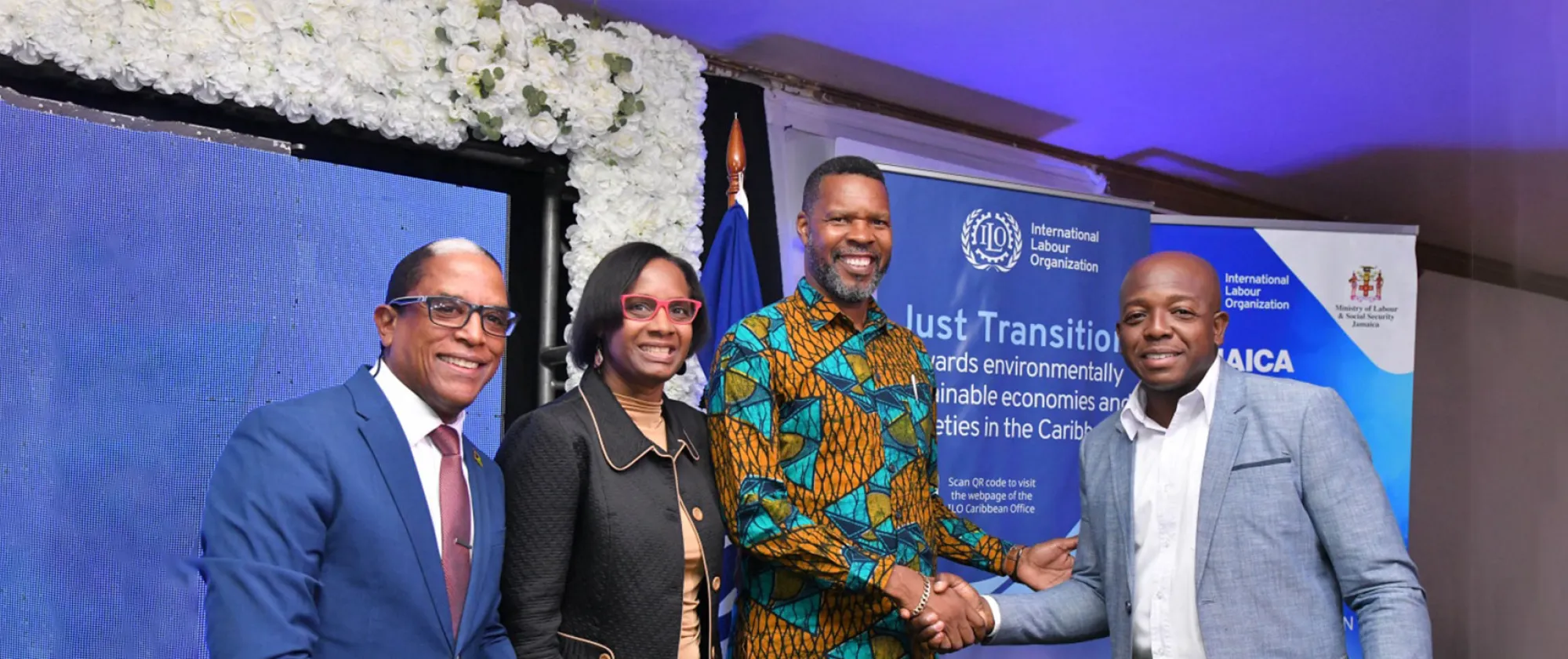 Four smiling people standing in front and a banner, with two of the men shaking hands