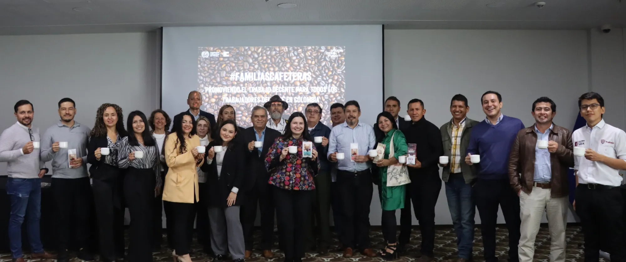 Un grupo de 23 personas posan para la foto con una taza de café en sus manos y tienen de fondo el letrero de la campaña #FamiliasCafeteras. En el centro de la fotografía, está Paola Campuzano, Coordinadora Nacional del Proyecto, quien sostiene además, una muestra de café molido producida por una cooperativa de café de paz.