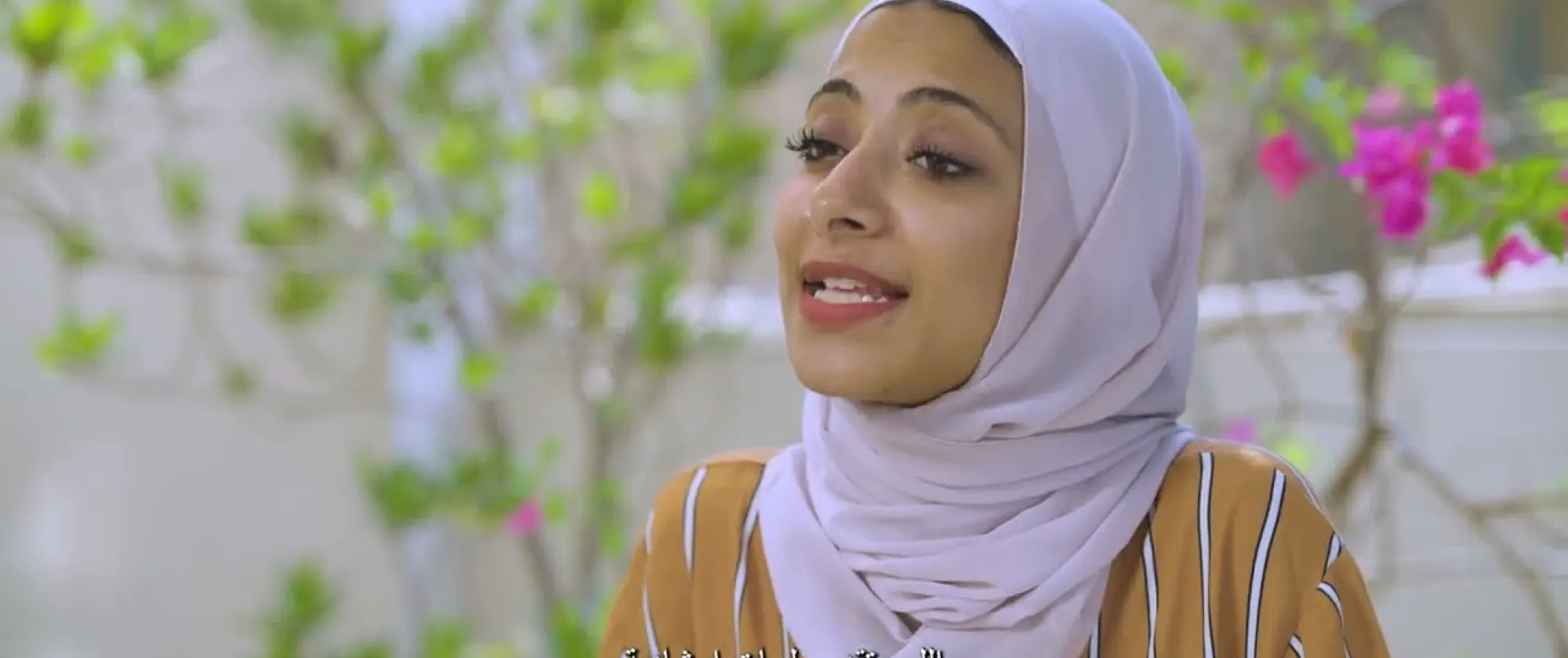 Woman in a hijab speaking outdoors, surrounded by green foliage and pink flowers, with subtitles in Arabic and English discussing guidance sessions.