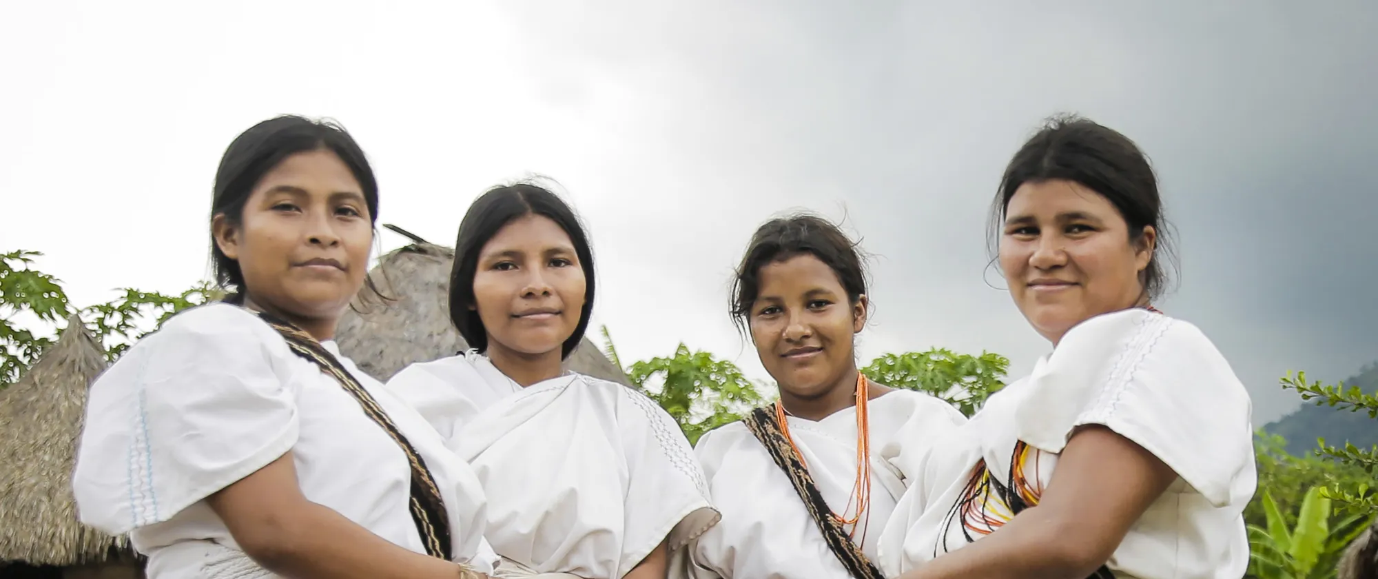 Cuatro mujeres indígenas luciendo su vestimenta tradicional con mantas de color blanco, collares y mochilas terciadas. De fondo una maloca indígena y naturaleza
