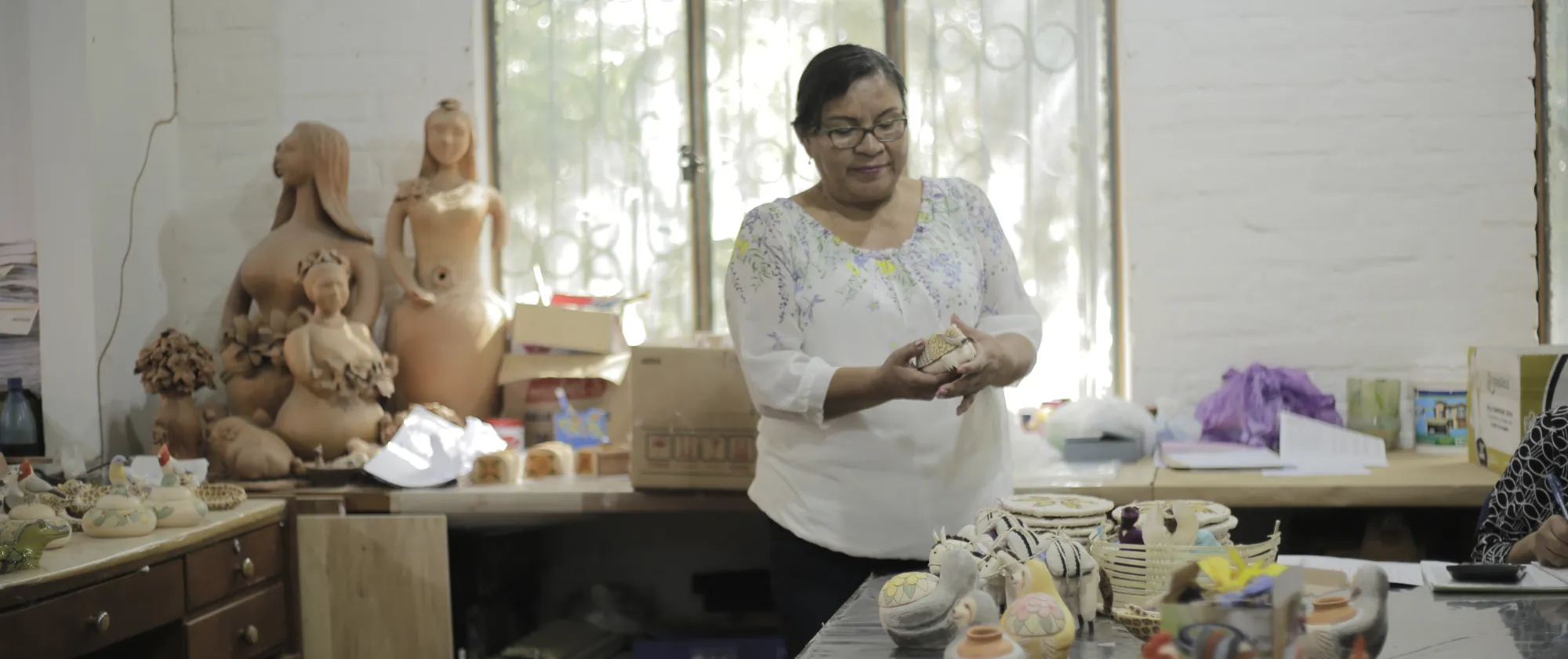 Mujer con lentes y camisa blanca en un taller de trabajo ordenando productos realizadas a base de cerámica, alrededor de ella diversas artesanías finalizadas como figuras femeninas y animales como burros y gallinas