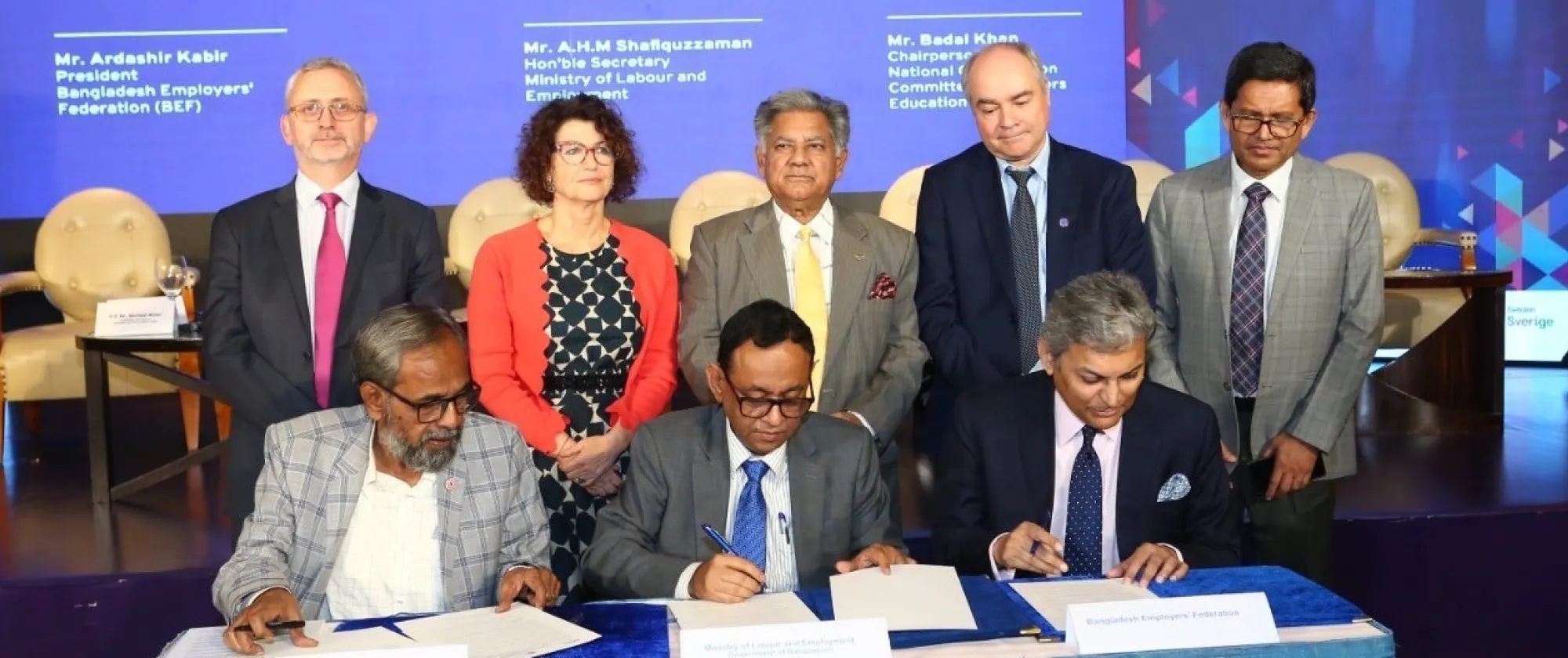Three men seated sign documents as other people who are standing watch them.