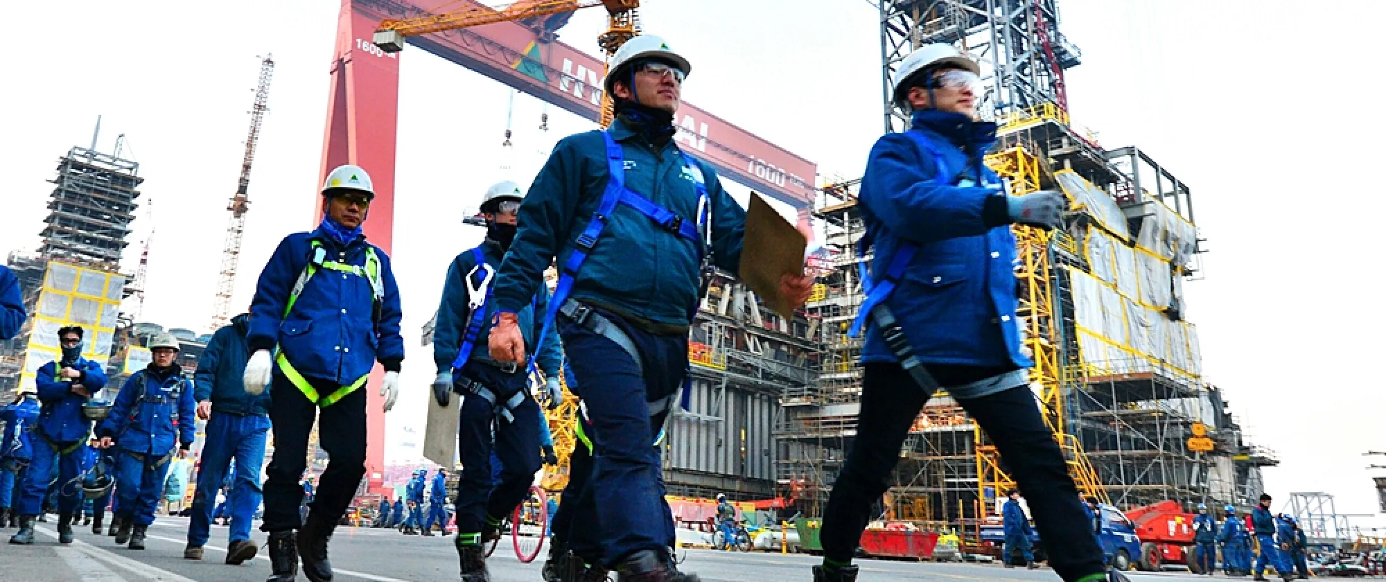 Workers wearing safety gear, including helmets and gloves, walking at a busy industrial site with cranes and construction equipment in the background.